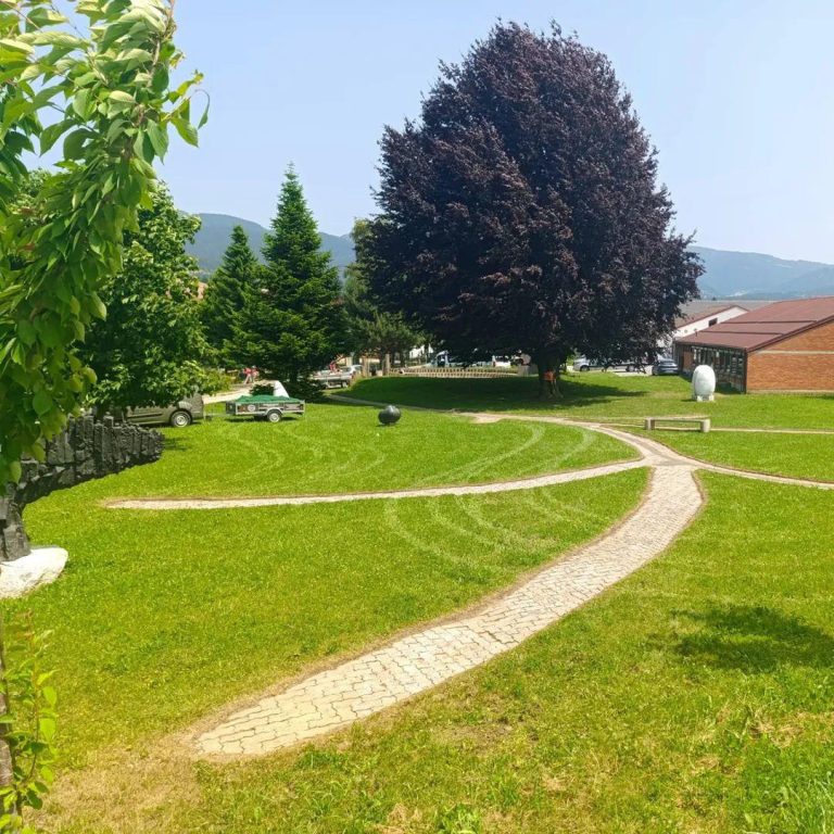 Pokošena parkovna površina z vijugastimi tlakovanimi potmi in vozilom Mojstra Jake v ozadju